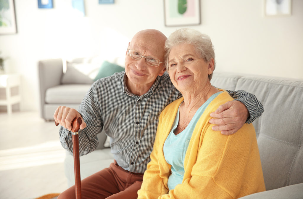 Senior couple sitting next to one another and hugging each other while looking at camera in senior home