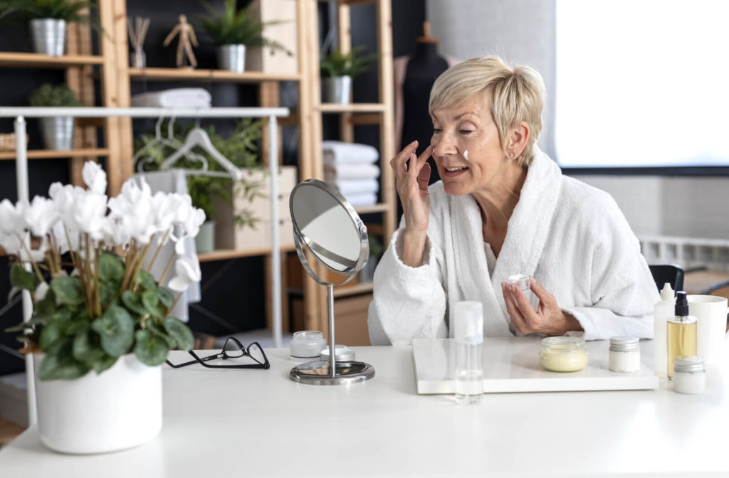 A mid-aged woman with short blonde hair in a white bathrobe sits at the table in front of a mirror and puts cream on her face inside the living room.