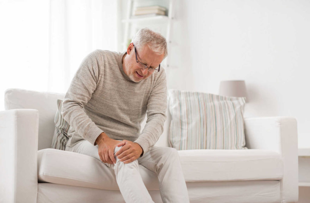 An elderly man sitting on a couch, holding his knee as he experiences severe joint pain.