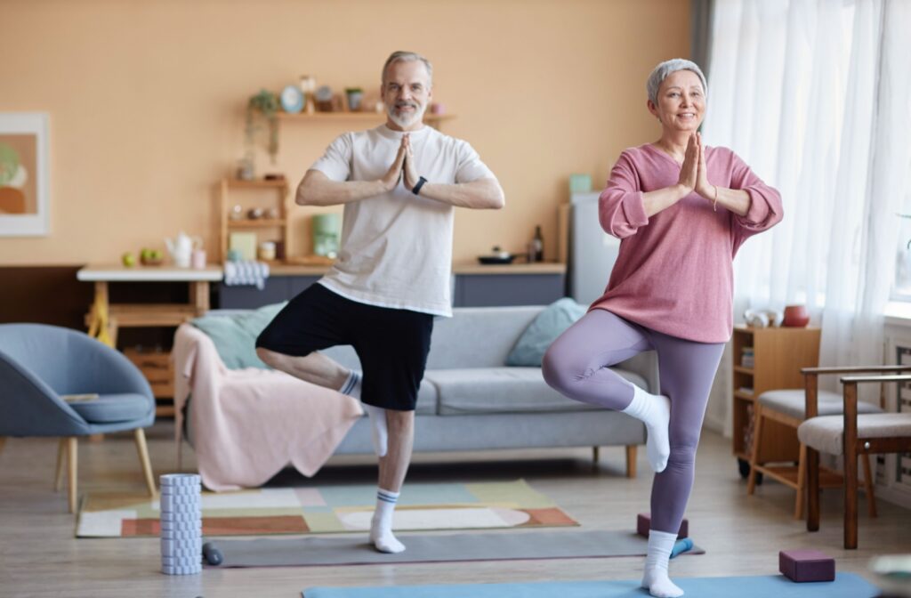 An older couple performs yoga tree poses together in a cozy living room