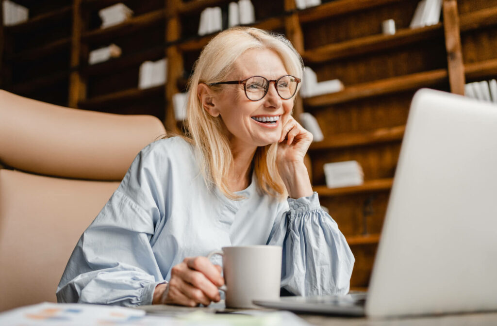 An older adult smiling over coffee while looking at an independent living community on their laptop.