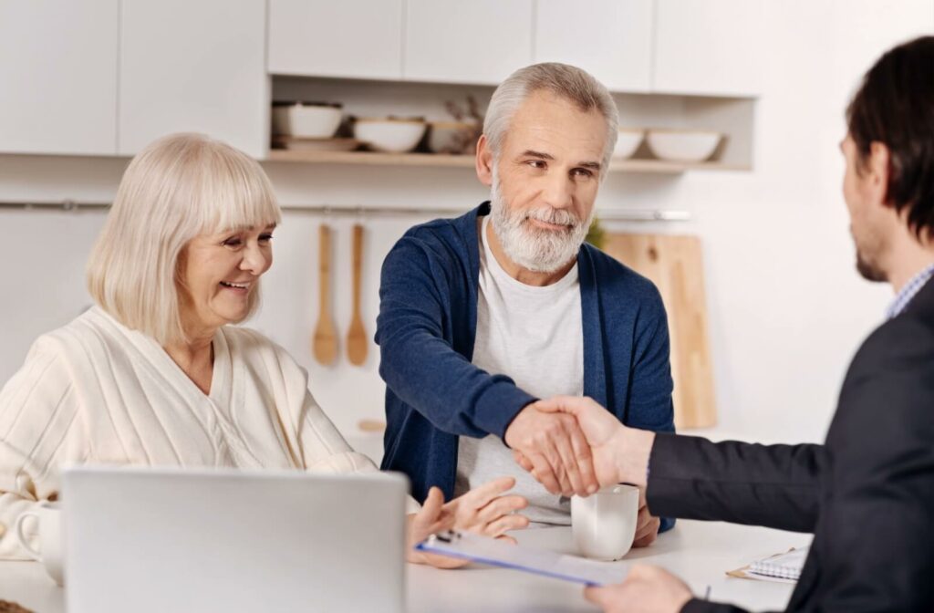 Older adult shaking hands with advisor across table while another person smiles during a retirement planning meeting in modern kitchen