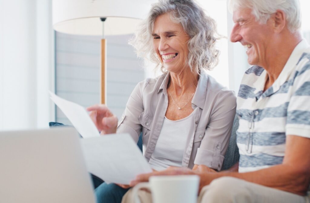 Two older adults laughing while reviewing retirement documents together at home with laptop and coffee